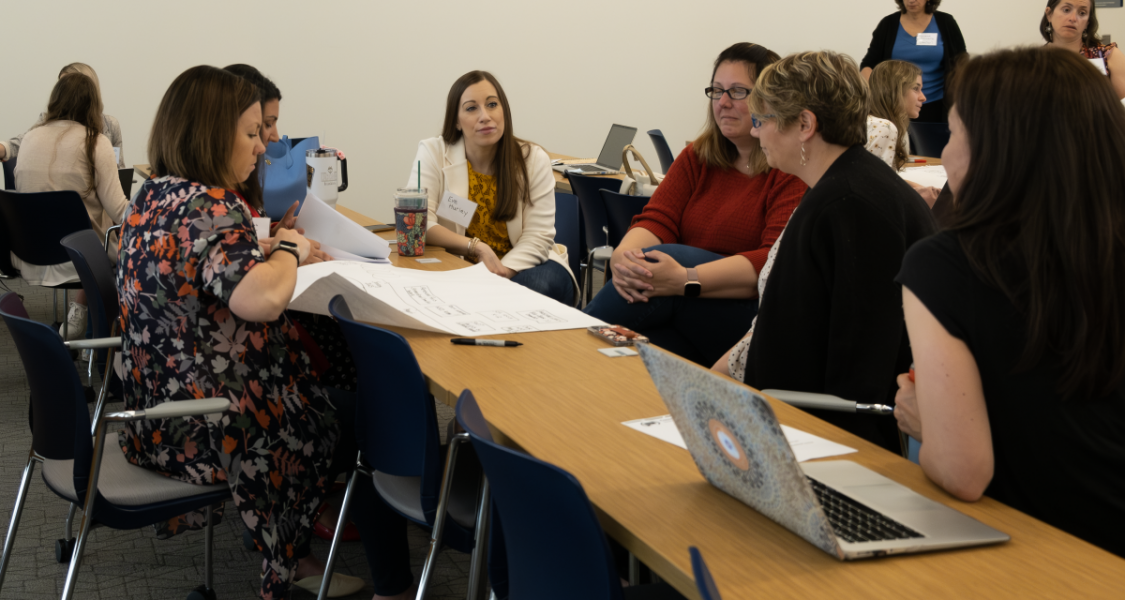 a small group of 6 people sit at long tables looking at a large sheet of paper