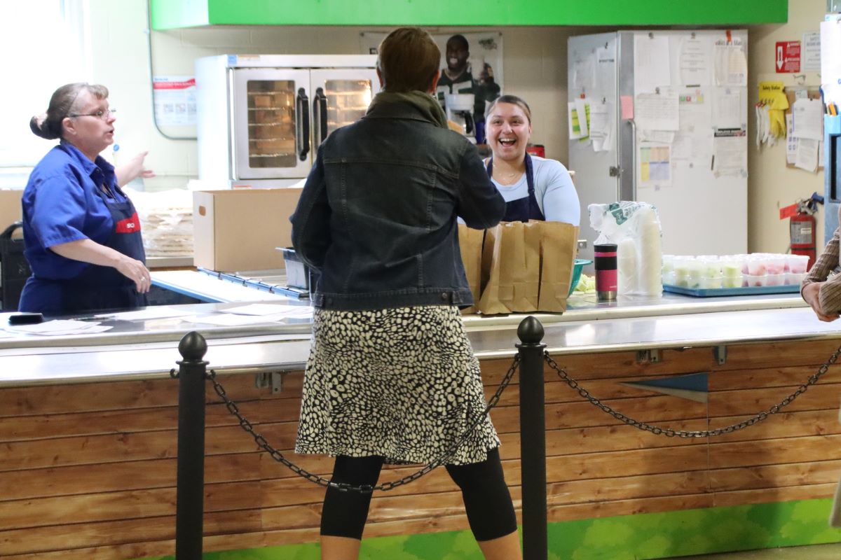 two women stand behind a cafeteria counter talking with a person standing in front with their back to the camera