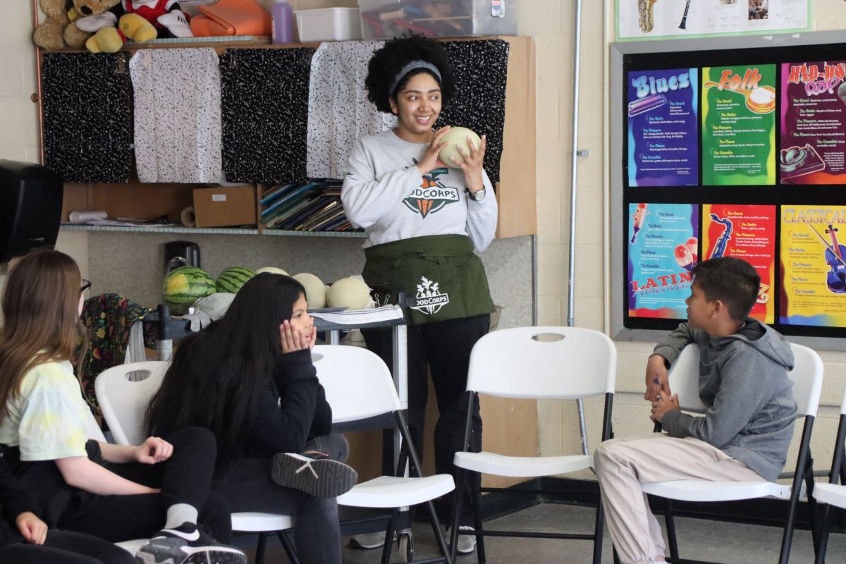 An adult stands smiling holding a honeydew lemon. Three children sit in chairs watching her