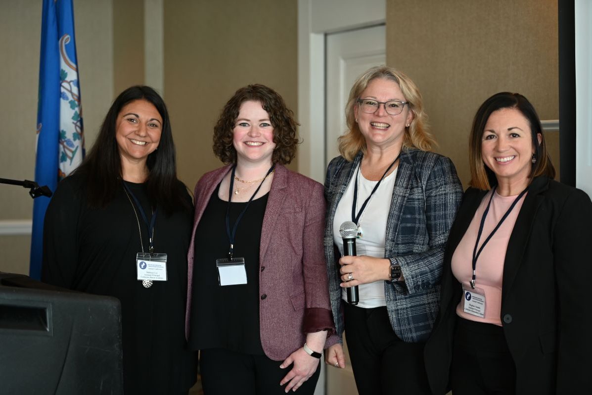Melissa Cyr, Kate Williamson, Sandra Chafouleas, and Meghan Amado stand and smile at the camera at a conference. Sandra Chafouleas is holding a microphone.