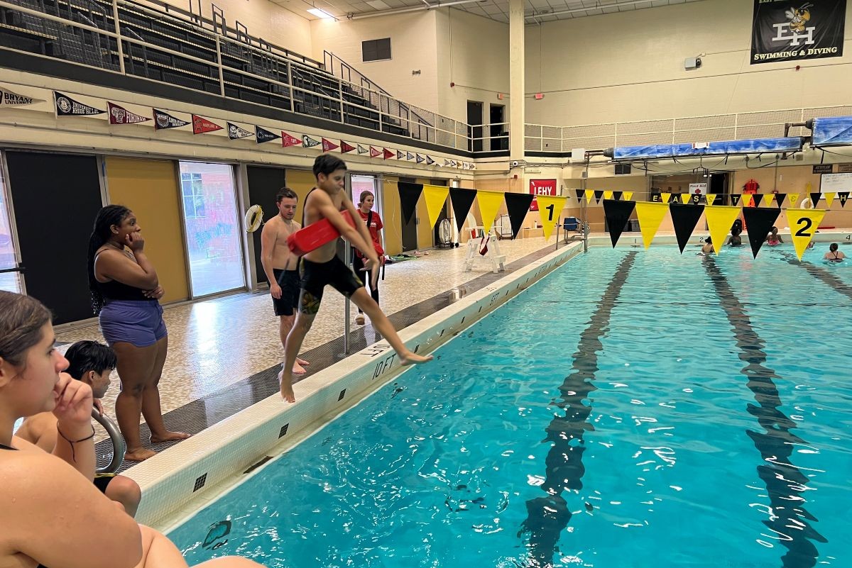 A child steps off the edge of a swimming pool, ready to jump in with other children looking on.