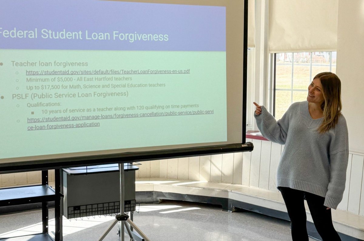 Woman presenting a slideshow in the front of a room. The title of the slide reads 